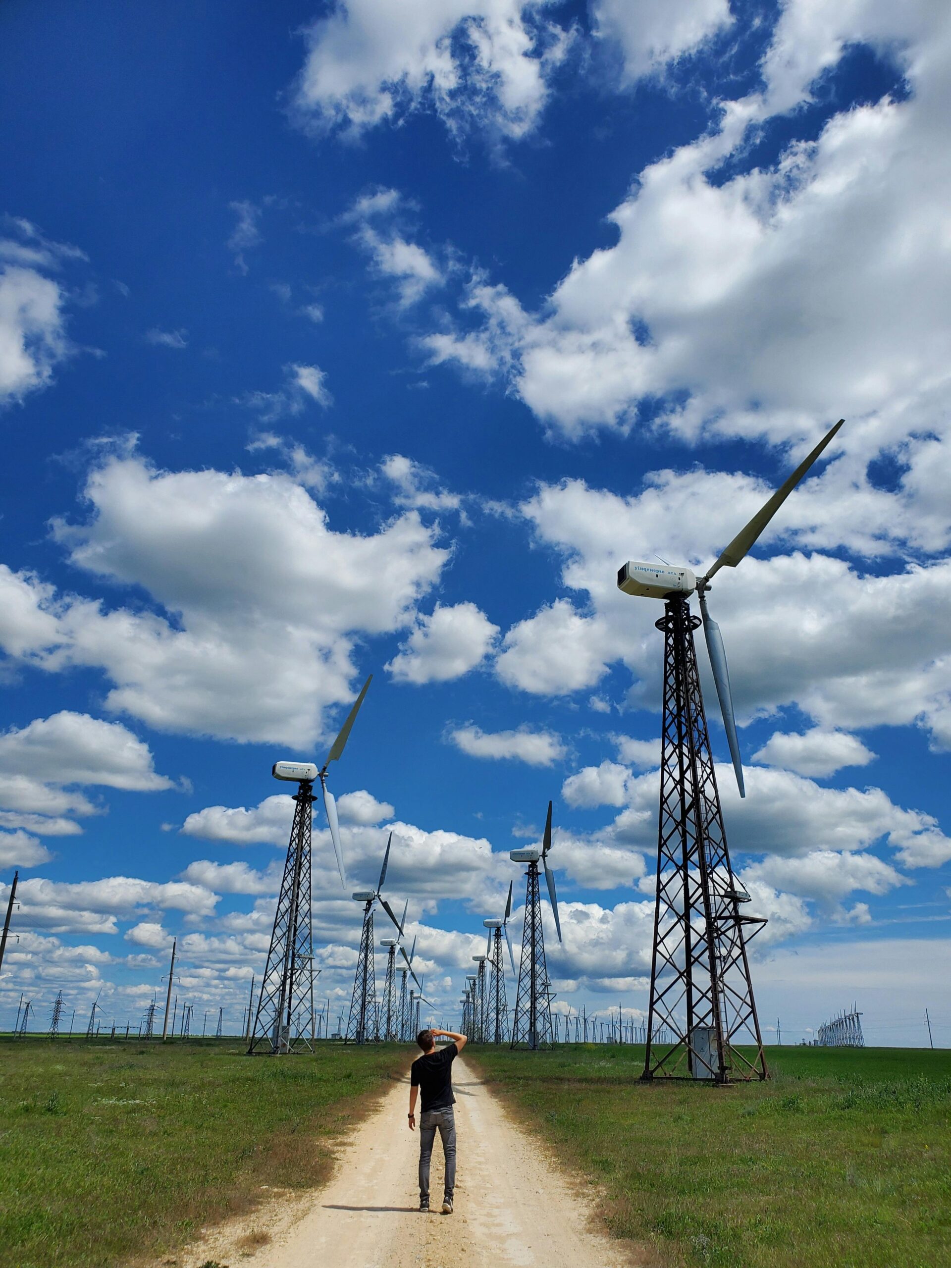 A man walks on a path surrounded by wind turbines under a blue sky with clouds, symbolizing renewable energy.